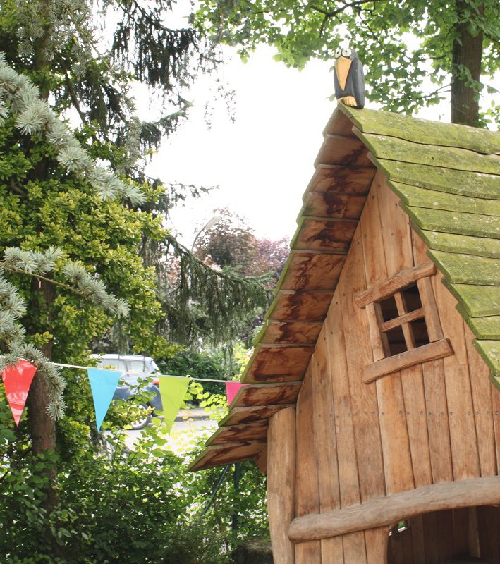 Spielhaus aus Holz mit einem grünem, bewachsenen Dach und bunten Fähnchen im Hintergrund.